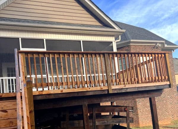 Wooden deck with railing attached to a house, featuring screen porch and blue sky background.