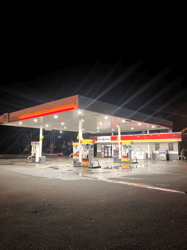 Shell gas station at night, illuminated pumps with wet pavement reflecting lights.