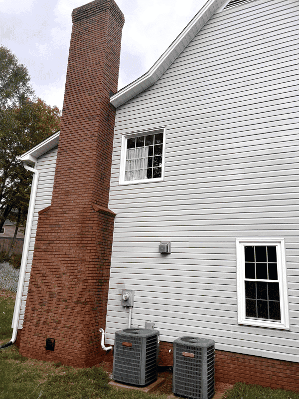 Back view of a house with a brick chimney and two outdoor HVAC units against gray siding.