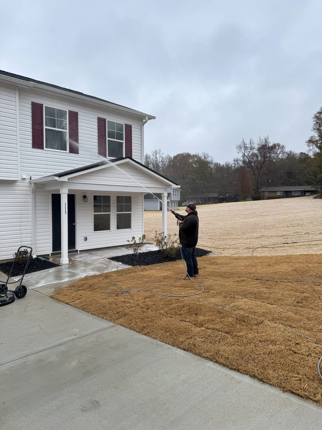 Man pressure washing a house exterior on a cloudy day, with newly laid grass nearby.