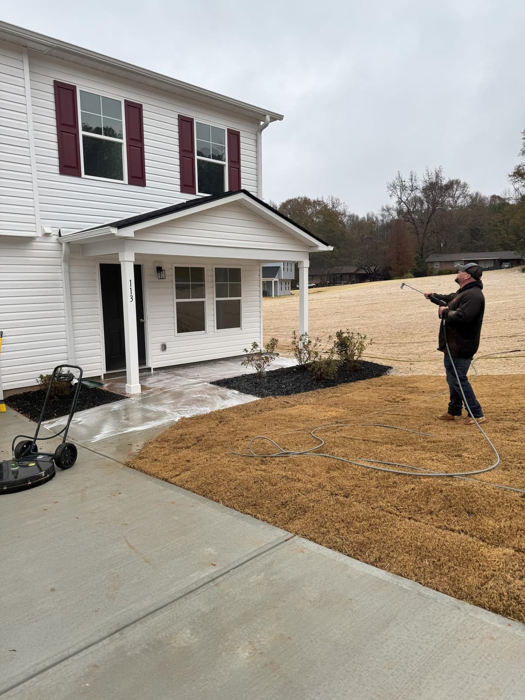 Man power washing a house exterior and driveway on a rainy day. New home in the background.