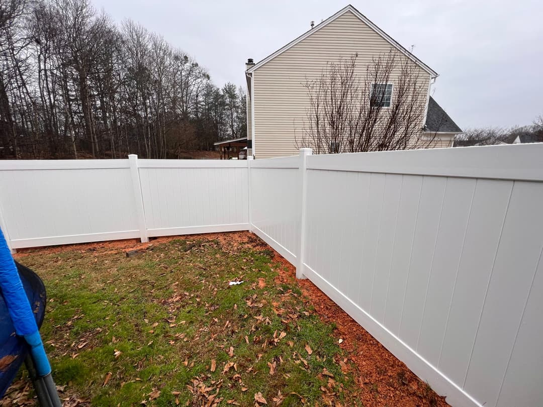 White vinyl fence enclosing a grassy yard, with trees in the background and a house nearby.