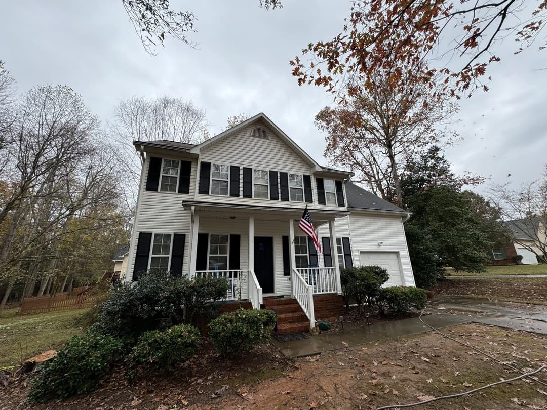 White two-story house with black shutters and an American flag in front, surrounded by trees.